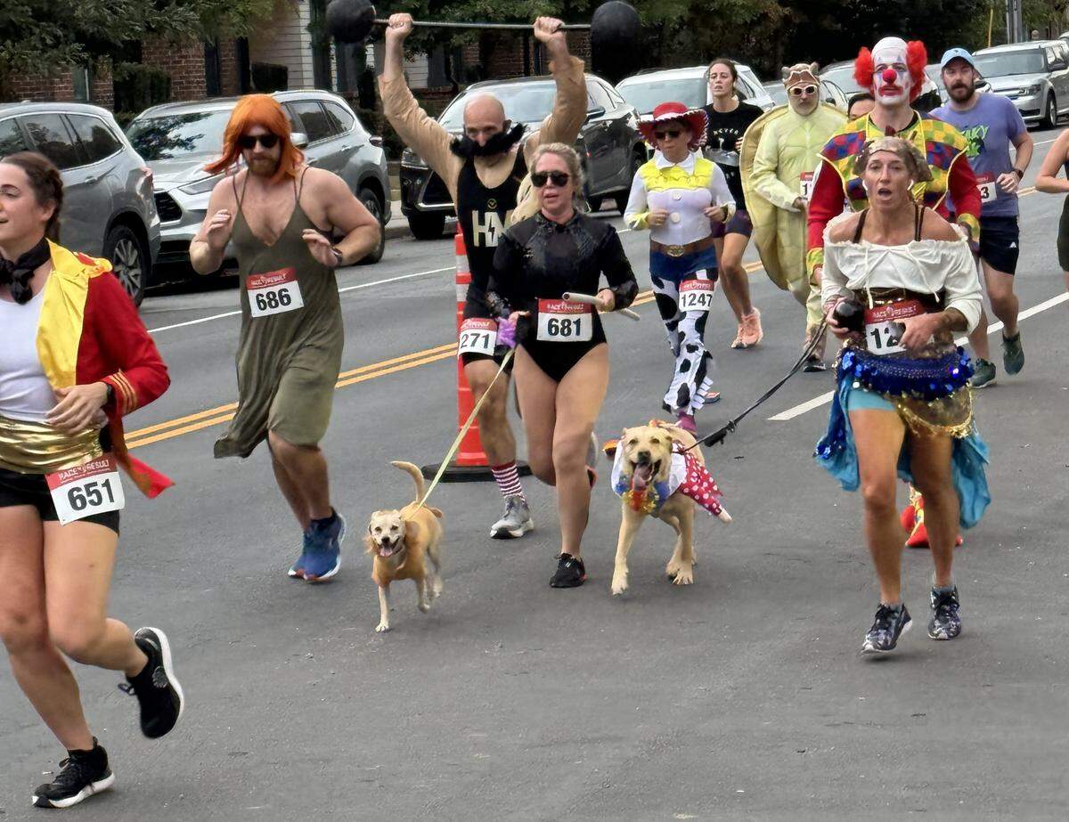 A dynamic, eye-level shot of a group of people participating in a costume race on a paved street. The runners are wearing a variety of costumes, including a ringmaster, a man in a green dress with a red wig, a circus strongman, a woman in a black leotard walking two dogs, Jessie from Toy Story, a scary clown, and a woman in a pirate wench costume. All participants, wearing race bibs, are in motion, running down the street.