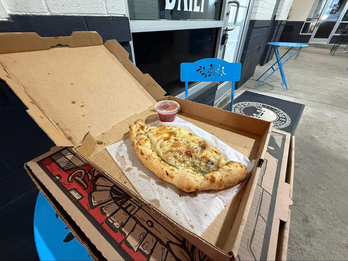 A large, herb-topped calzone served with a side of marinara sauce sits in an open pizza box on an outdoor table.