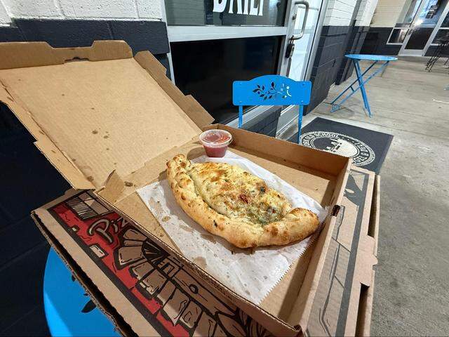 A large, herb-topped calzone served with a side of marinara sauce sits in an open pizza box on an outdoor table.
