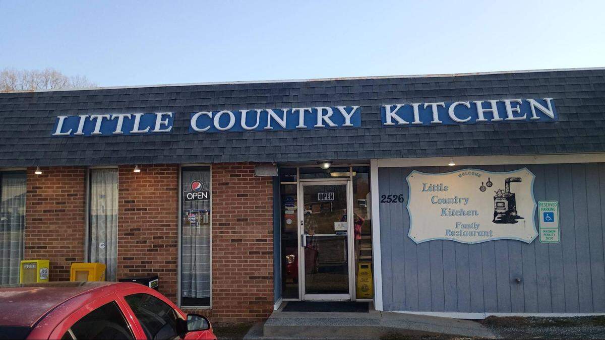 A building features a dark shingled mansard-style roof overhang with large blue block letters that read “LITTLE COUNTRY KITCHEN.” To the right of the glass entrance door, a decorative white sign reads “Welcome Little Country Kitchen Family Restaurant” next to an illustration of an old-fashioned wood-burning stove.
