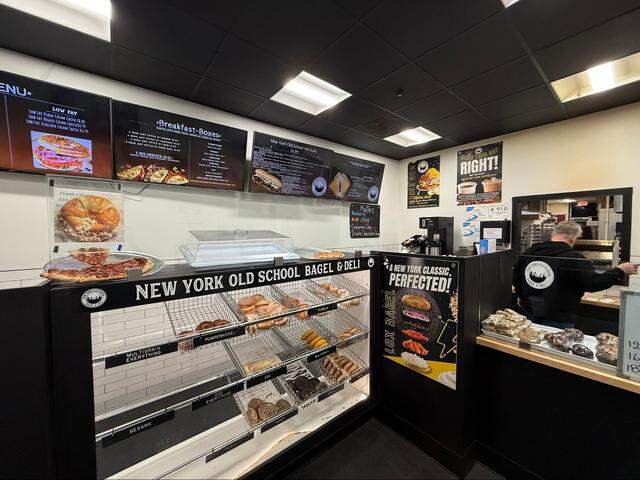 An indoor, eye-level shot shows the service counter of “NY Old School Bagel & Deli.” A large glass display case in the foreground is filled with various bagels organized in wire baskets, labeled with varieties like Multigrain Everything, Pumpernickel, Sesame, and Poppy. Above the case sits a tray of sliced pepperoni pizza.