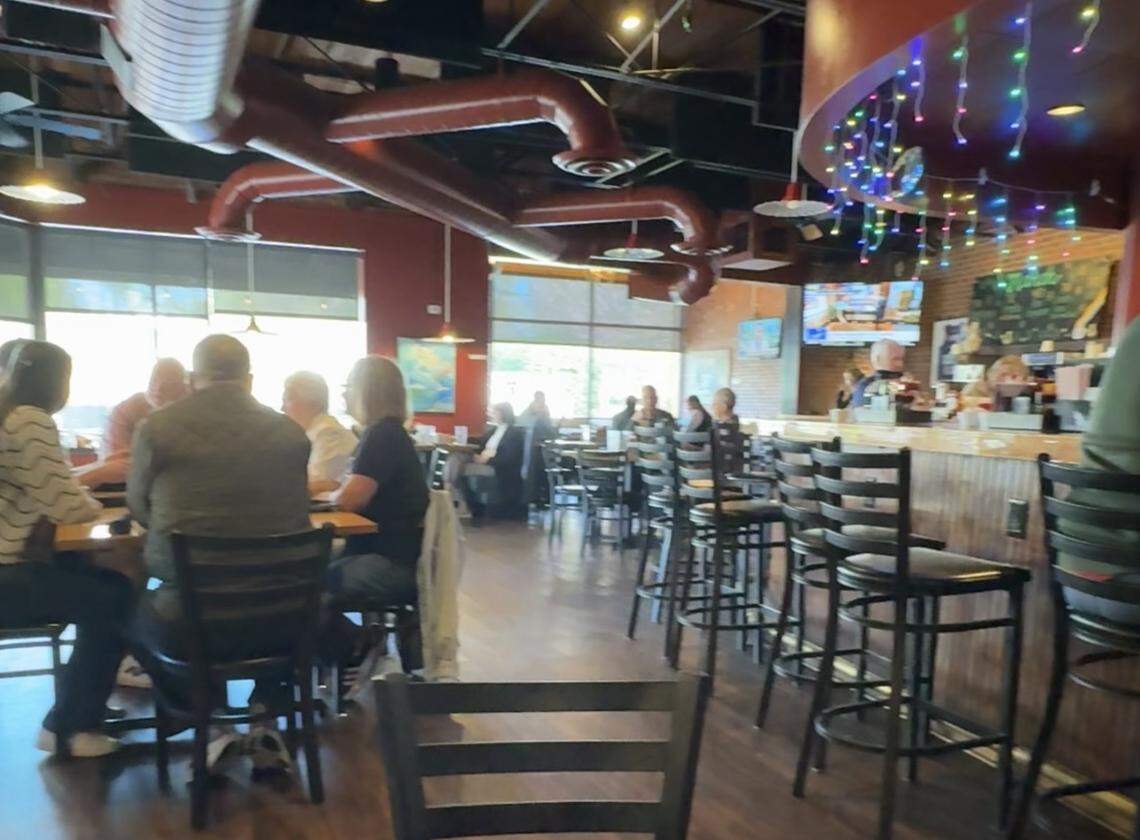 A bustling morning scene inside a casual, neighborhood-style diner. The restaurant features a warm atmosphere with dark wood floors, exposed red ductwork along the ceiling, and large windows that fill the space with natural light.