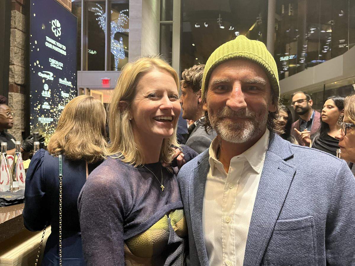 A close-up shot of a couple posing together at a crowded indoor event. The person on the left with blonde hair is smiling broadly. The other, in the foreground, has a beard and is wearing a blue blazer and a lime green beanie, looking at the camera. In the background, other people are mingling and a blue banner thanking partners is visible.