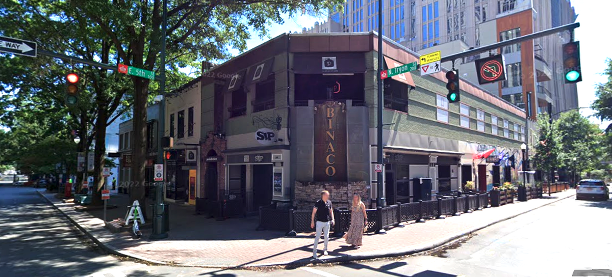 A two-story corner building with a bar and restaurant on the ground floor is visible from a street intersection. Two people are walking on the sidewalk in front of the building, which has a sign that reads “BINACo.”
