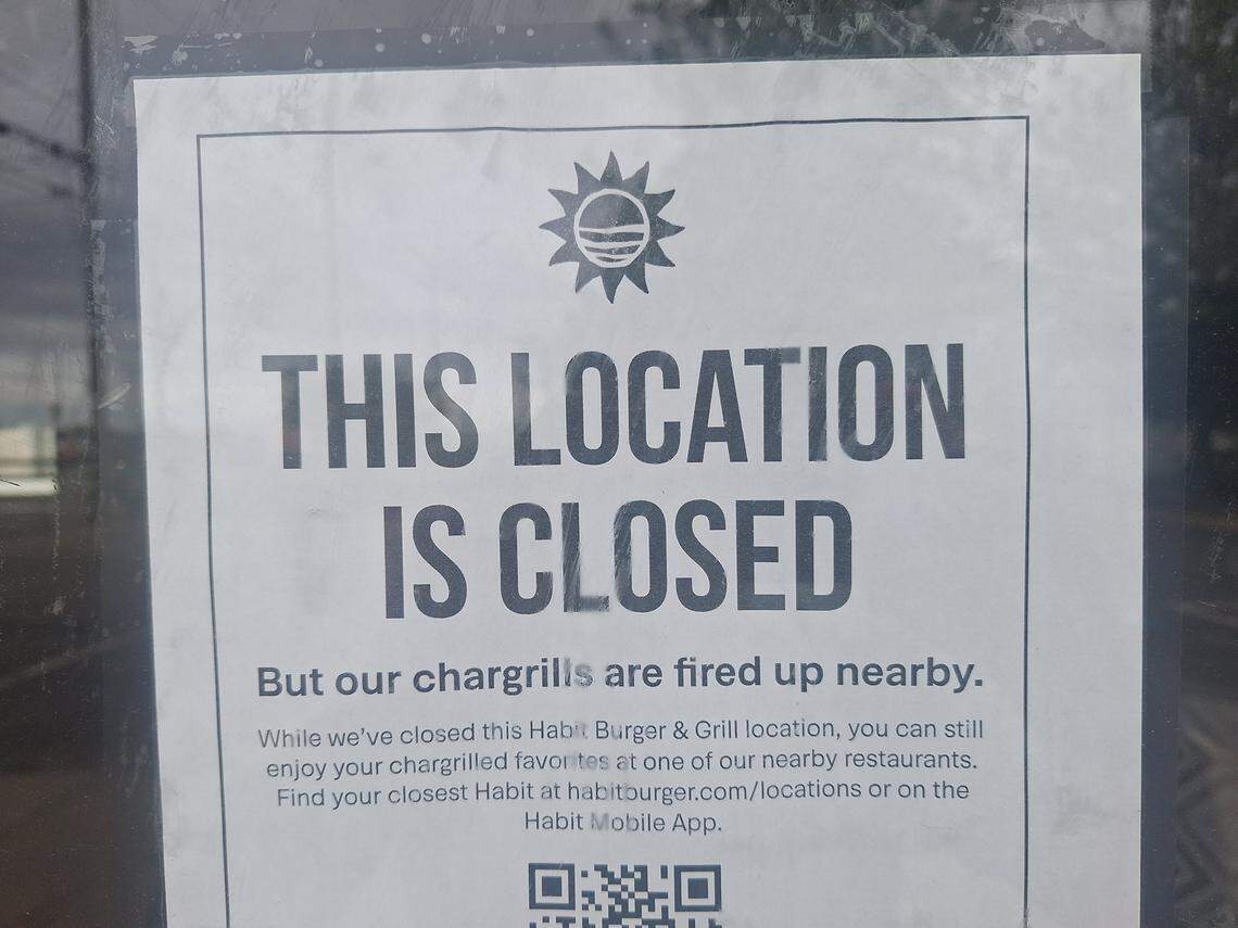 A black and white sign posted on a glass door features a sun logo and large text stating, “THIS LOCATION IS CLOSED.” Below the main heading, smaller text explains that while this Habit Burger & Grill has closed, customers can find nearby restaurants via their website or mobile app. A QR code is partially visible at the bottom of the notice to help patrons locate the next closest chargrill.
