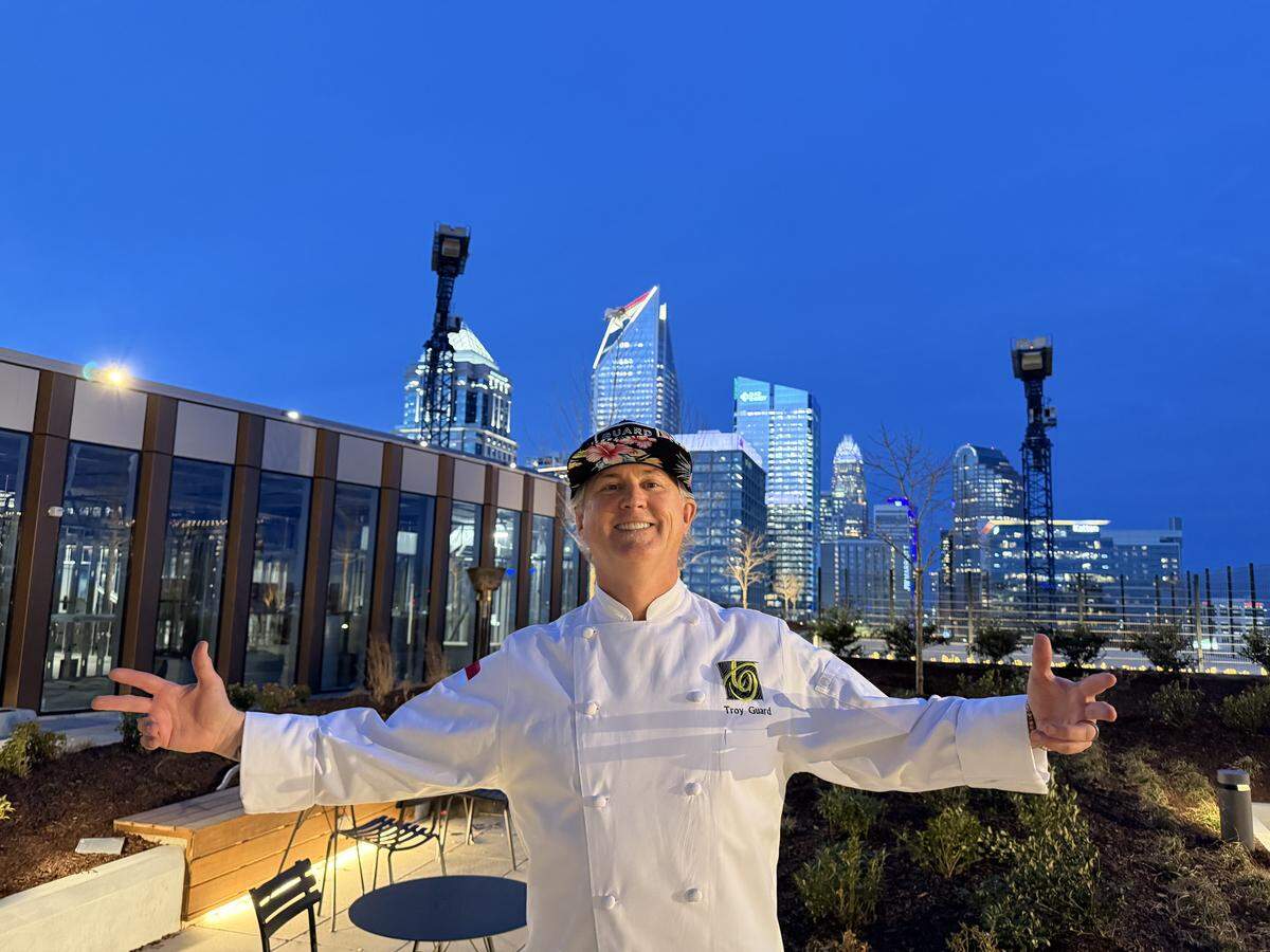 A medium shot of a chef standing on a rooftop patio at night with his arms outstretched in an inviting gesture and the illuminated skyline of Charlotte, North Carolina, in the background. The chef is wearing a professional white chef’s coat with “Troy Guard” embroidered on the chest and a black baseball cap featuring a colorful floral pattern on the brim. The background features several recognizable skyscrapers from the Charlotte skyline, including the distinctive curved top of the Truist Center and the lit-up Duke Energy building. The sky is a deep twilight blue.