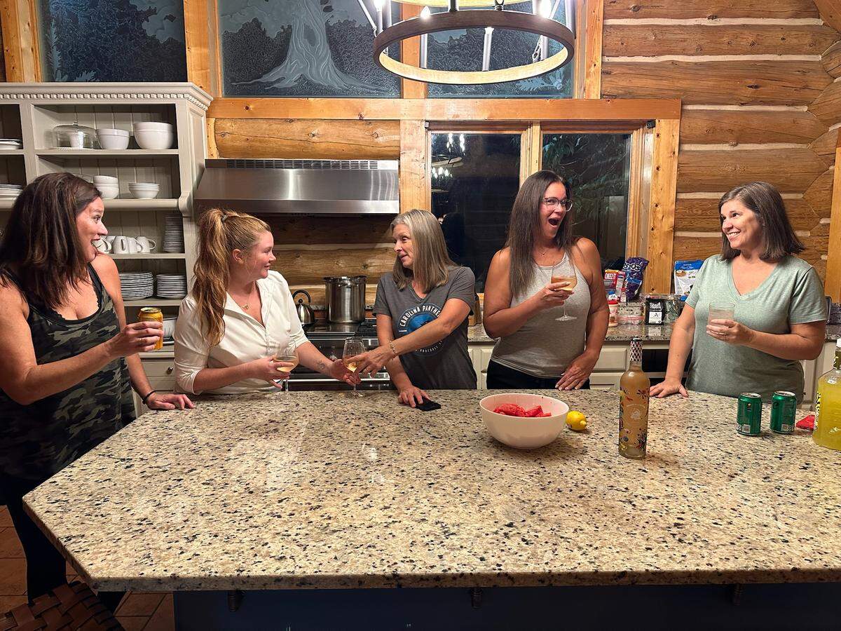 A group of five women enjoys a social gathering in a log cabin-style kitchen. They are standing around a large granite countertop, holding wine glasses and other drinks, and are engaged in lively conversation and laughter.
