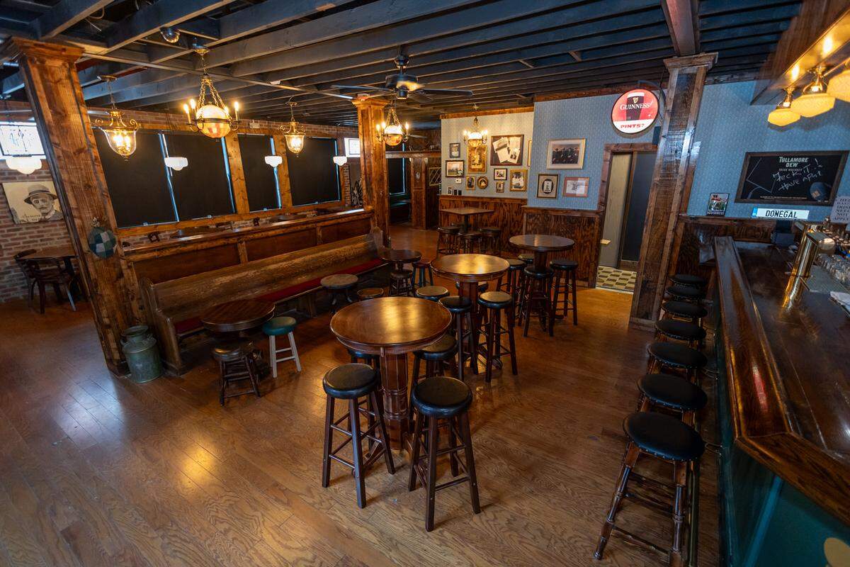 A wide-angle interior view of a traditional Irish pub with warm wooden floors and dark exposed-beam ceilings. The room features high-top circular tables with black cushioned stools, a long wooden bar on the right, and a unique vintage church-pew bench along a central partition. The walls are decorated with framed historical photos, a glowing Guinness sign, and blue patterned wallpaper above wood paneling. Ornate brass lanterns hang from the ceiling, casting a warm, inviting glow throughout the empty tavern.