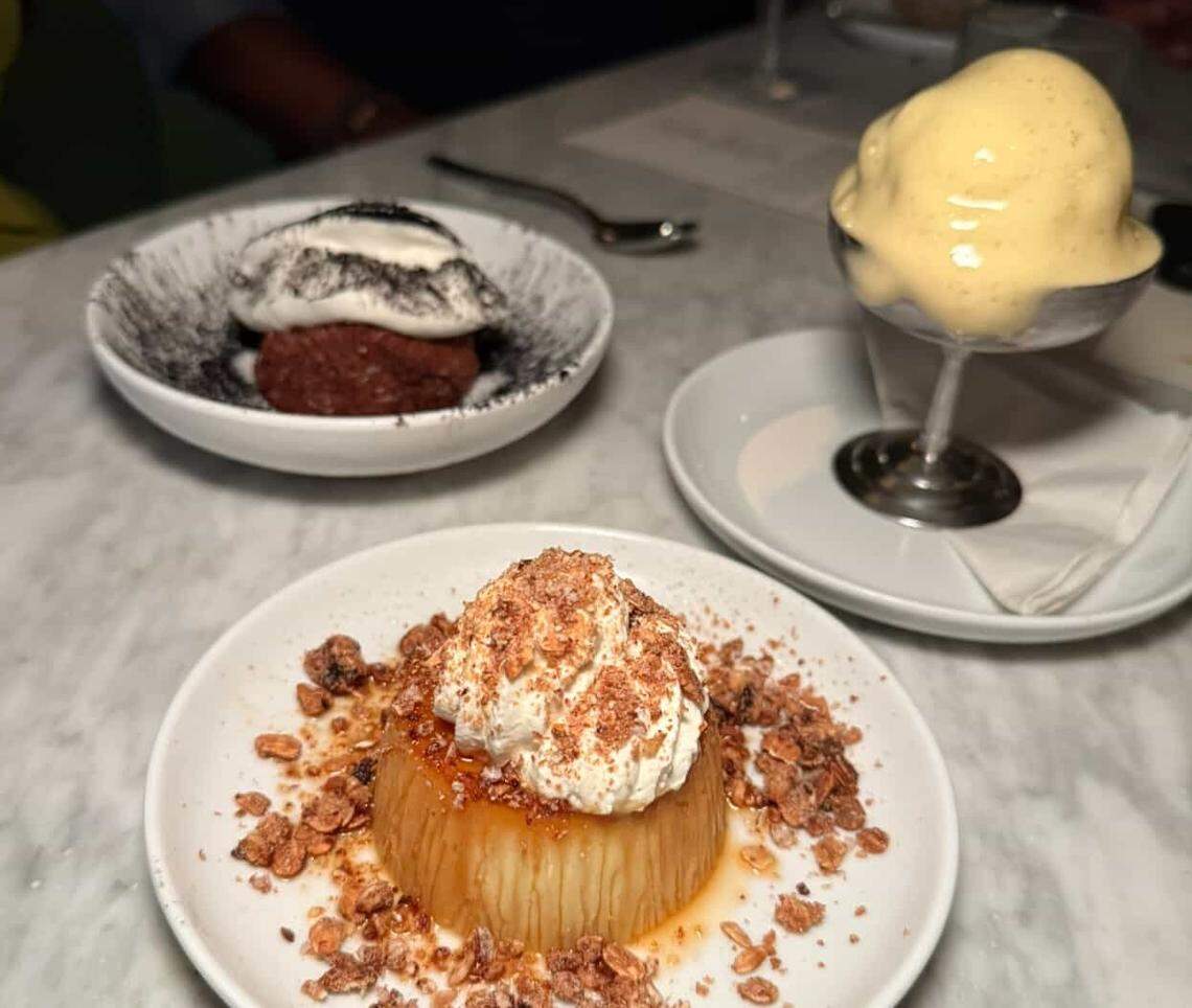 A trio of desserts from a Rada and Customshop collaboration are arranged on a white marble table. In the foreground, a round, flan-like dessert with a light caramel sauce is topped with a dollop of white cream and a generous sprinkle of brown crumbles, possibly nuts or cookies. To the left, a small bowl with a speckled rim holds a dark red cake-like dessert with a contrasting mound of white cream and a dark powder garnish. To the right, a stemmed silver cup holds a large scoop of a pale yellow, airy dessert, possibly mousse or ice cream, with a subtle drizzle of sauce. The desserts are presented on small white plates.