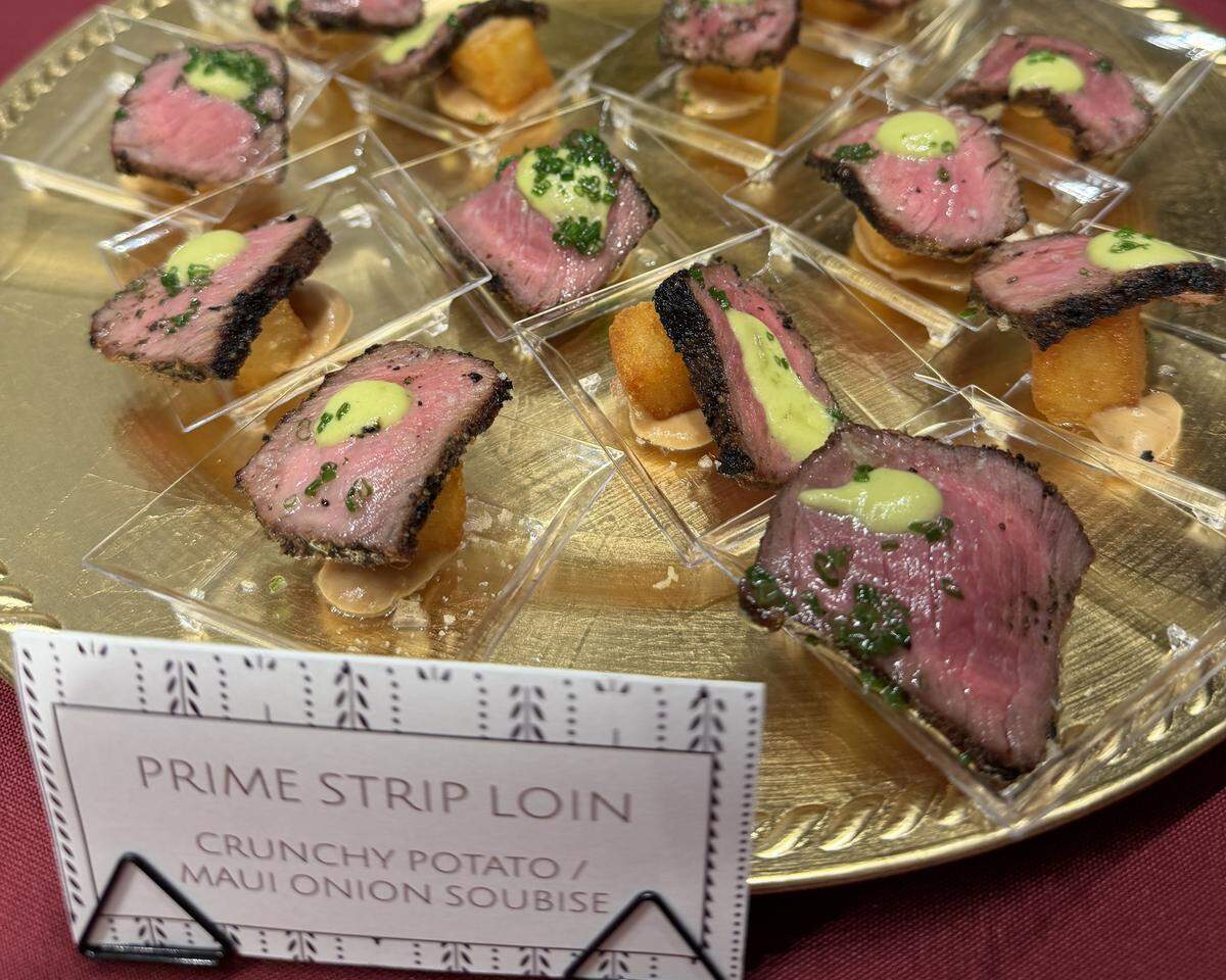 Individual portions of Prime Strip Loin are served medium-rare with a vibrant green herb oil and a dollop of creamy sauce. Each slice of beef is perched atop a crunchy potato bite and a Maui onion soubise, as noted by the display card in the foreground.