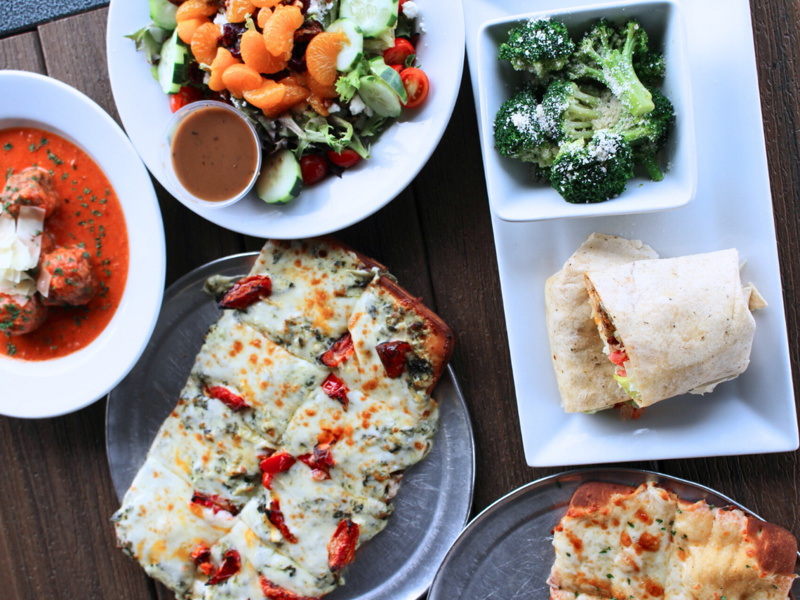A table full of different dishes including a plate of meatballs in red sauce, a side salad with mandarin oranges, a bowl of steamed broccoli with grated cheese, a flatbread pizza with melted cheese and red peppers, and a turkey wrap.