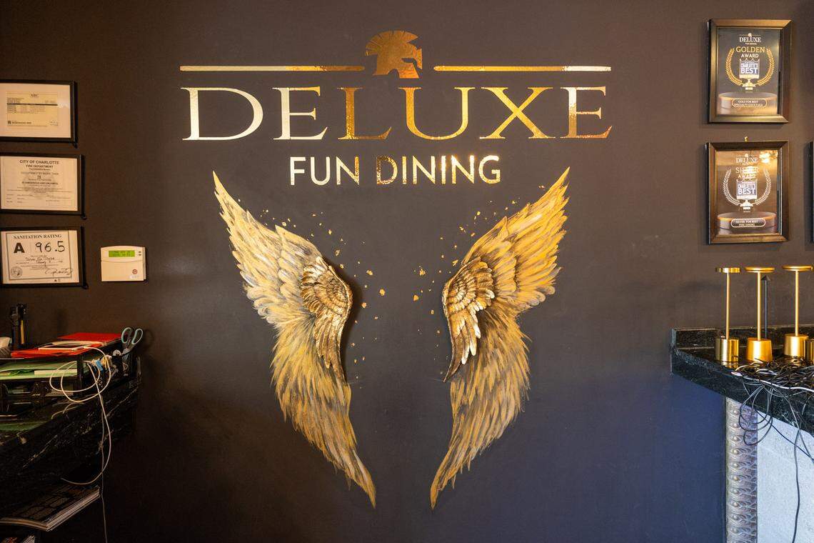 A black wall in a restaurant with gold lettering that says “DELUXE FUN DINING” and a silhouette of a Spartan helmet. Below the text is a pair of gold, feathered angel wings painted on the wall. To the left are several framed awards and certificates, including a City of Charlotte inspection rating of 96.5. To the right are more framed awards and a marble countertop.