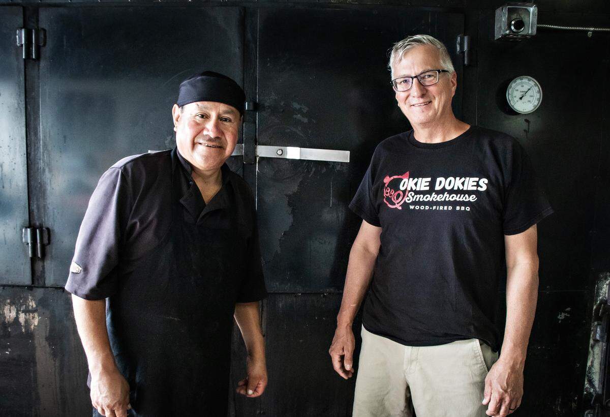 A well-lit portrait of two smiling men. José, on the left, is a Hispanic pitmaster in a black uniform and cap. Steve Dunning, on the right, wears glasses and a logo t-shirt for his restaurant, Okie Dokies Smokehouse. They are standing in front of the heavy-duty doors of their large, black, wood-fired smoker.