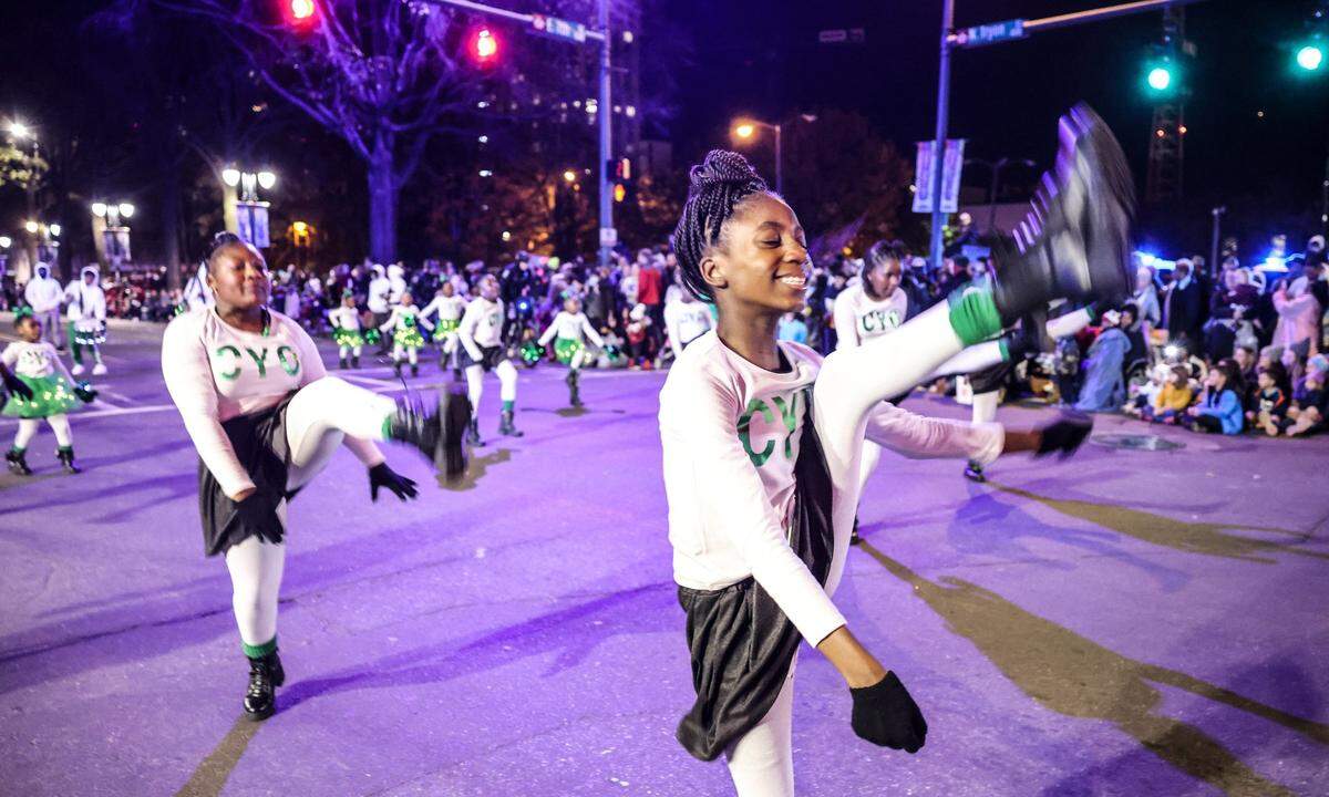 Two young female dancers in white shirts, black skirts, and white leggings perform a high kick motion in the foreground of a night parade. The street is illuminated by purple stage lights and background streetlights, showing a larger group of similarly dressed performers and spectators lined along the sidewalk.