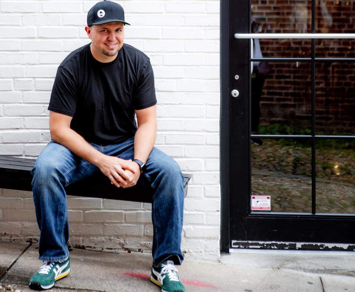 A person wearing a black t-shirt, blue jeans, green sneakers and a black baseball cap sits on a black wooden bench, leaning forward with hands clasped. The person is positioned in front of a white brick wall and a black-framed glass door that reflects a red brick building across the street.