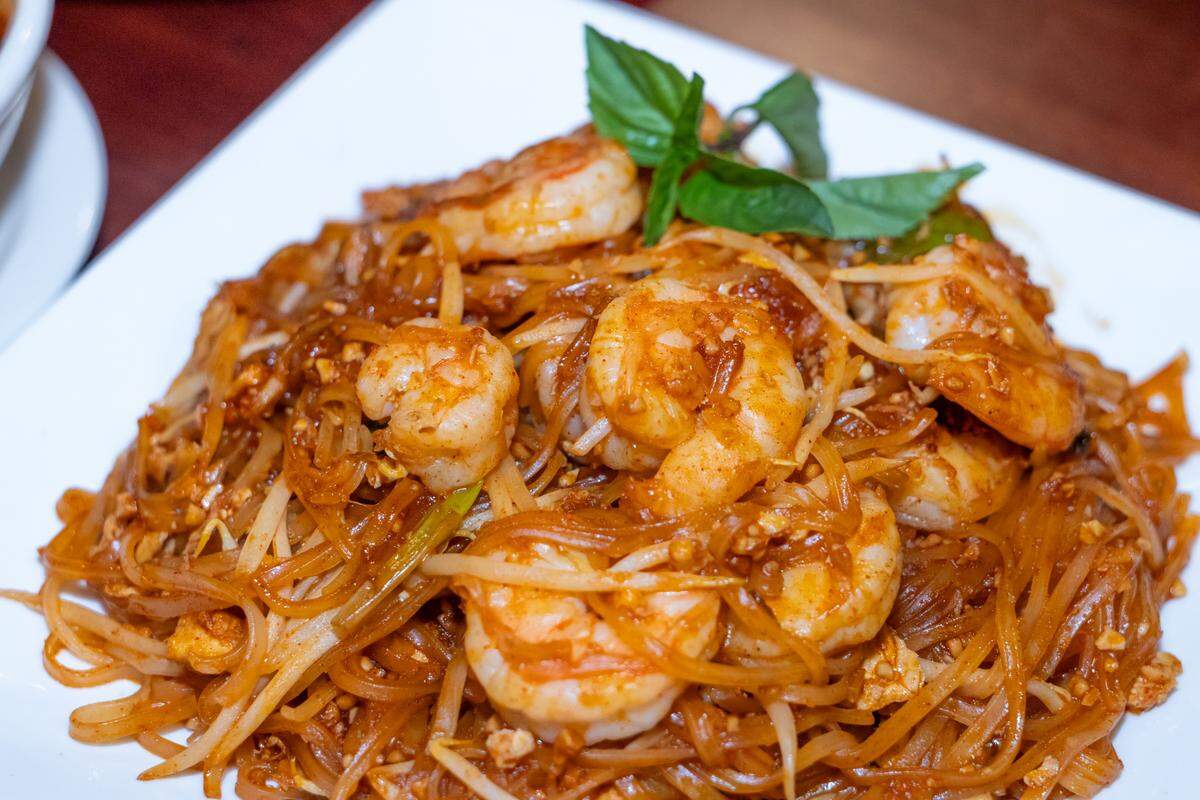 A tight close-up of a plate of shrimp Pad Thai. The dish shows thick rice noodles tossed in a reddish-brown tamarind sauce with large succulent shrimp, bean sprouts, and a garnish of fresh green basil.