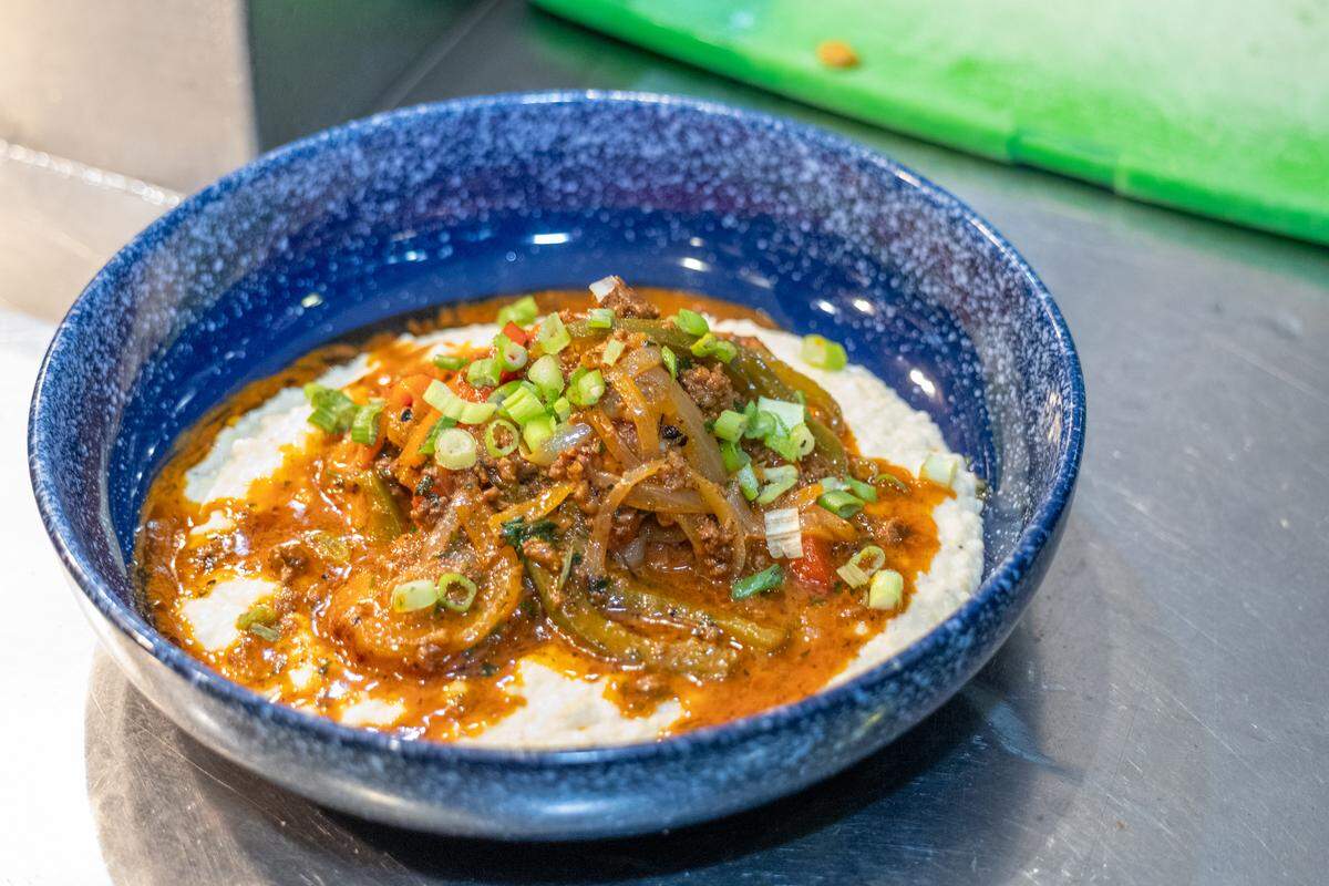 A top-down view of a blue speckled bowl containing a bed of creamy white grits topped with a savory meat and vegetable sauté. The dish is finished with a reddish sauce and a generous garnish of sliced green onions.