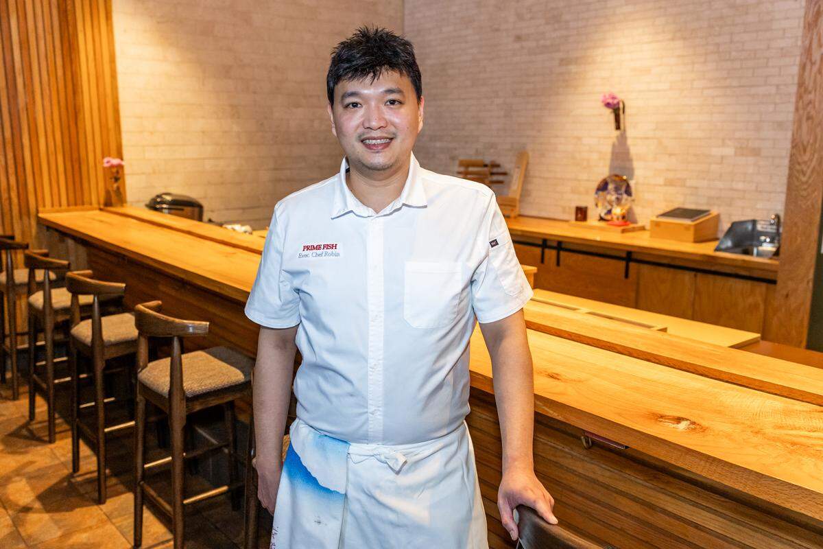 A chef poses behind a long, light-colored wooden sushi bar lined with high-backed wooden stools while smiling at the camera, wearing a clean white chef’s shirt and a traditional white apron.
