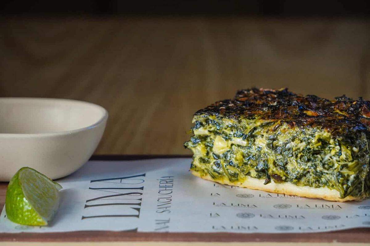 A close-up, eye-level shot shows a thick square slice of a savory green vegetable tart, possibly spinach or chard, with a golden crust and charred top. It is served on paper branded “LIMA ARTISANAL SANGUCHERIA” next to a small lime wedge and a white bowl.