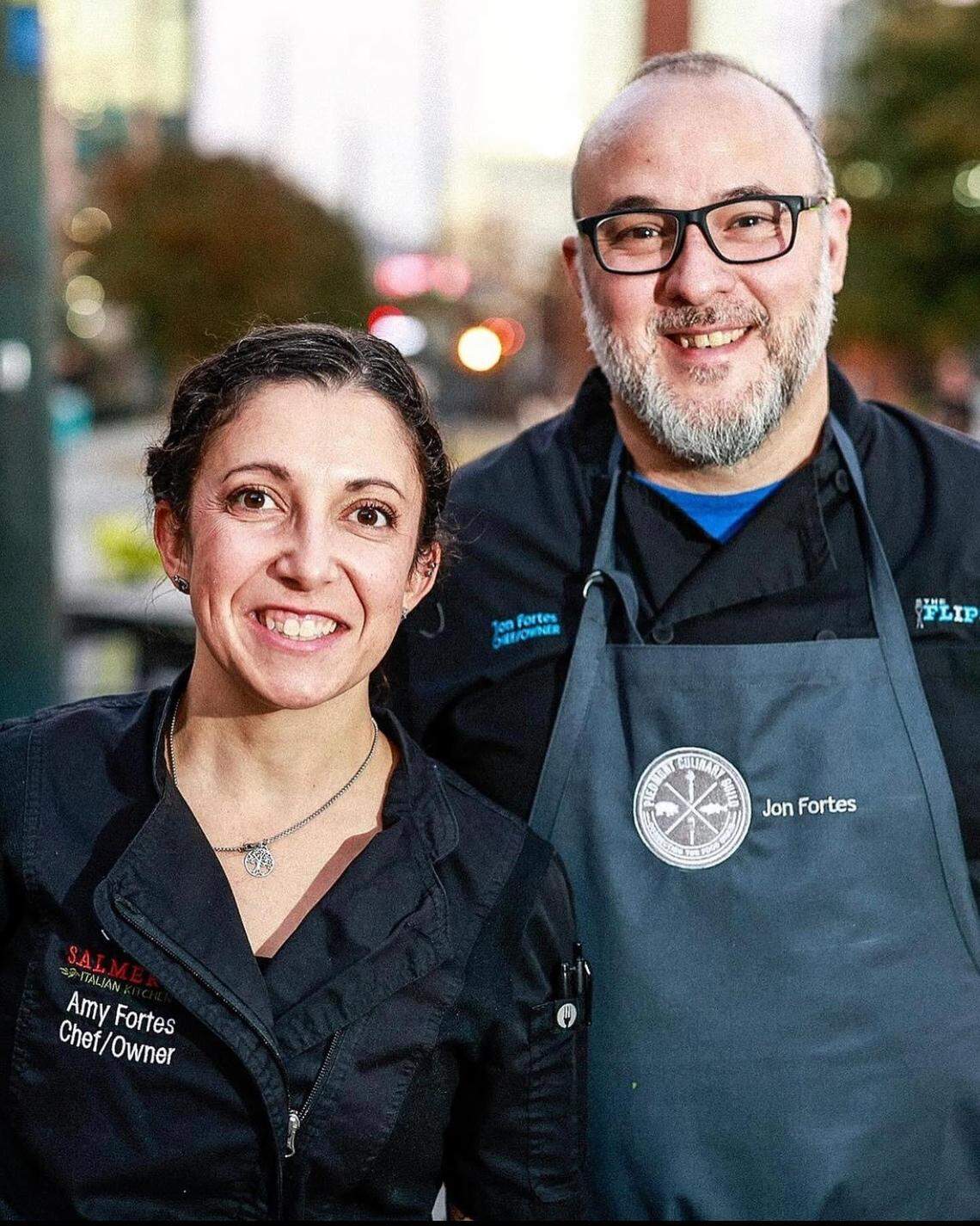 A portrait-style photograph of a man and a woman, who appear to be a couple, smiling at the camera. They are wearing black chef’s jackets. The woman on the left, Amy Fortes, has her hair pulled back and is wearing a silver necklace. Her jacket has “SALMO” in red text and “Amy Fortes Chef/Owner” in white text. The man on the right, Jon Fortes, has a beard and is wearing glasses. He has a dark gray apron over his jacket with a circular logo and “Jon Fortes” embroidered on it. The background is slightly out of focus with city lights visible.