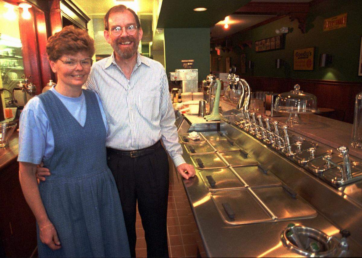 A 1994 photograph of a man and woman smiling behind the counter of a retro-themed ice cream parlor. The man wears a striped button-down shirt and glasses, while the woman wears a blue denim jumper. They are standing behind a long, polished stainless steel soda fountain counter equipped with vintage-style syrup pumps and ice cream containers.