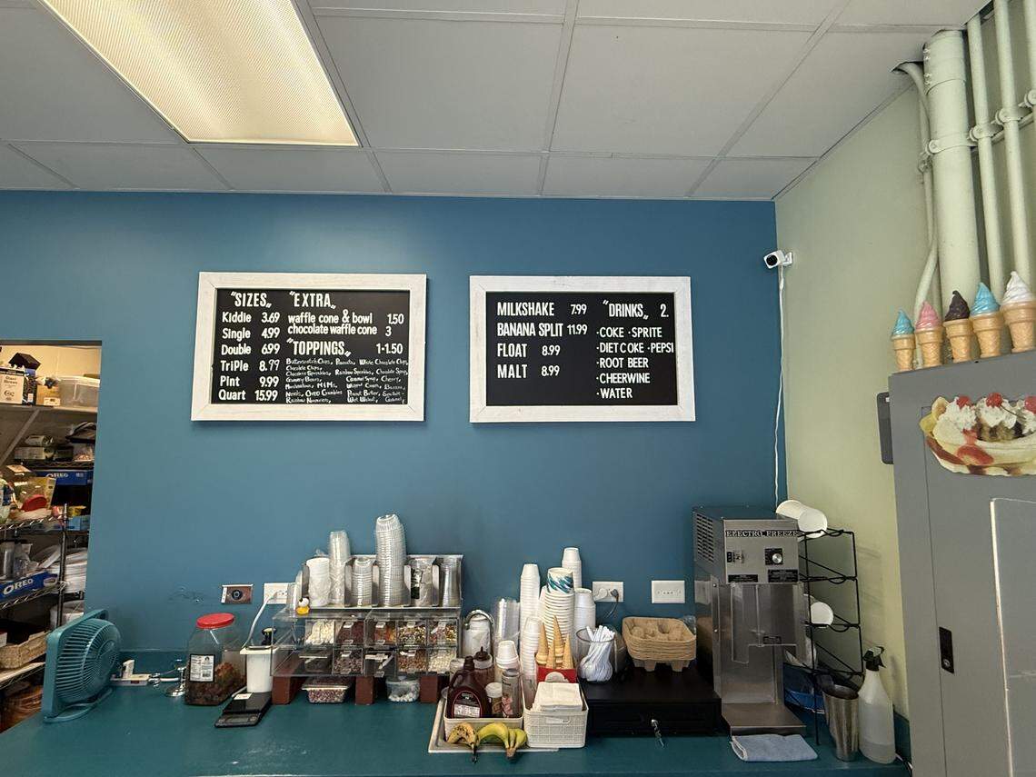 The interior service counter of the ice cream shop, featuring blue walls, two black menu boards with white text listing sizes and toppings, and various sundae supplies.
