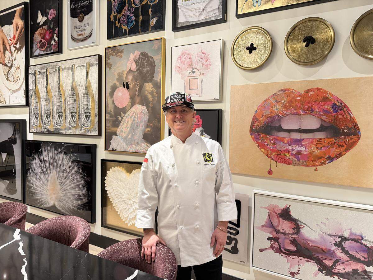 A chef is positioned at the end of a long, black-and-white marbled table, smiling and wearing a white chef’s coat with a name and a green logo embroidered on the chest, paired with a black baseball cap that features a floral pattern on the brim. Behind the chef is a floor-to-ceiling gallery wall filled with diverse contemporary art. The collection includes various styles, such as a large pop-art collage of lips, a woman blowing a pink bubble with gum, abstract floral paintings, and large decorative brass buttons.