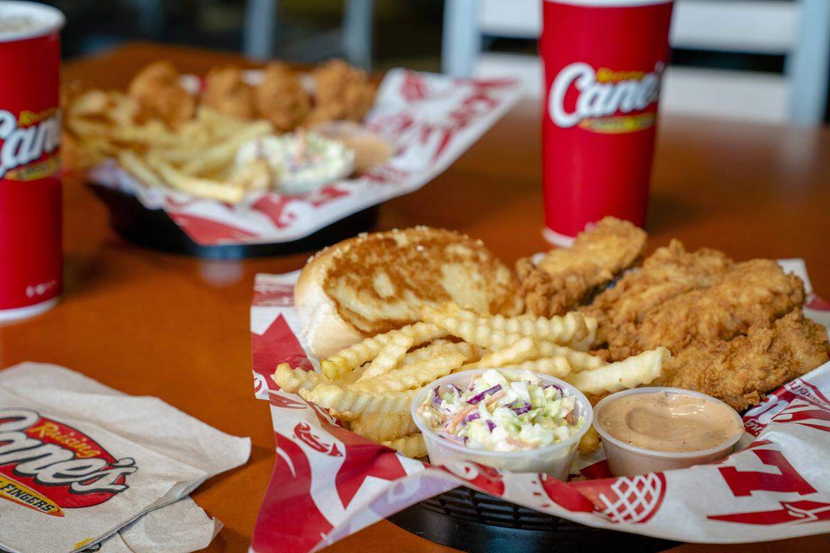 A warm, vibrant photo of a fast-food meal set on an orange table with black baskets lined with red-and-white branded paper hold fried chicken fingers, crinkle-cut fries, a slice of Texas toast, and a small cup of coleslaw.