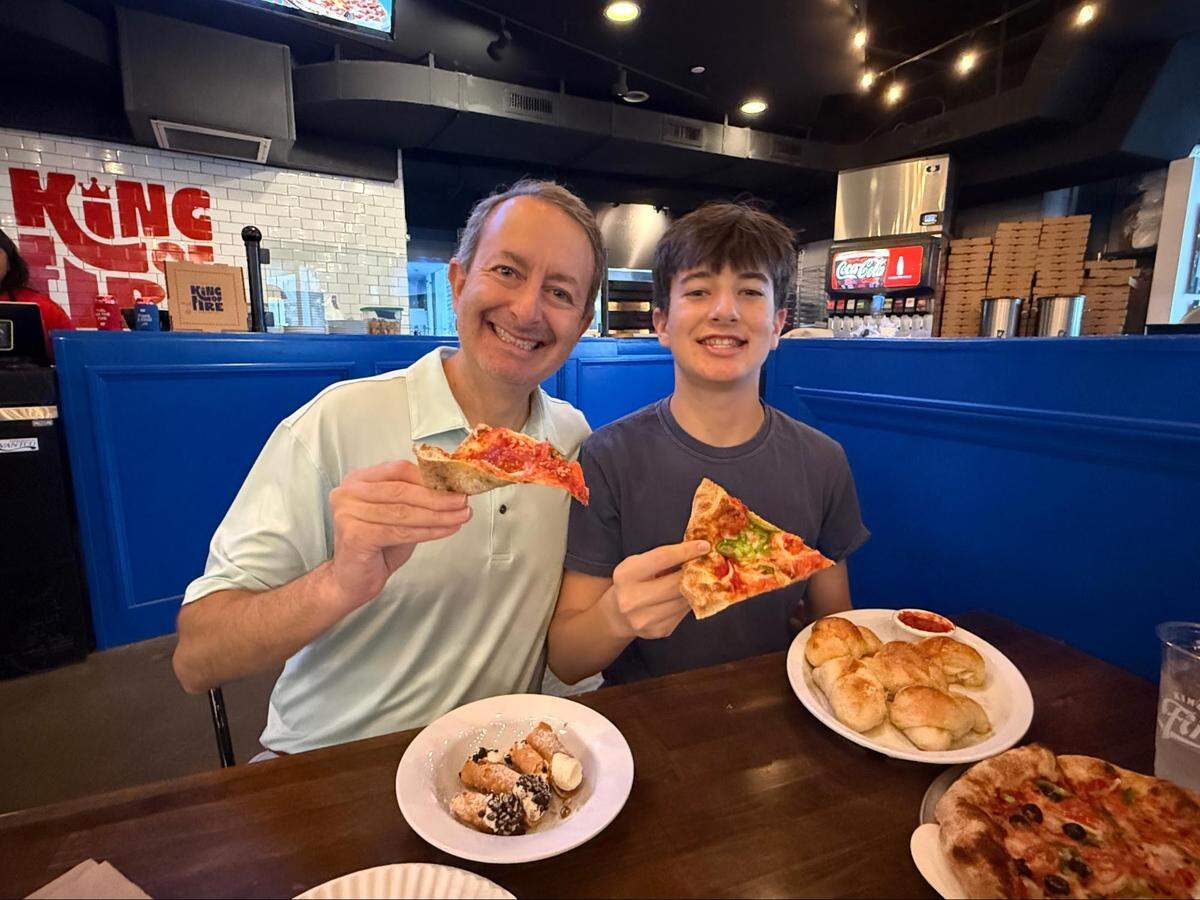 A person in a light green polo shirt and a younger person in the blue t-shirt sit side-by-side, each smiling and holding up a triangular slice of pizza. Their table is spread with garlic knots, a veggie pizza, and a plate of cannolis. The restaurant’s “King of Fire” branding is visible in the background.