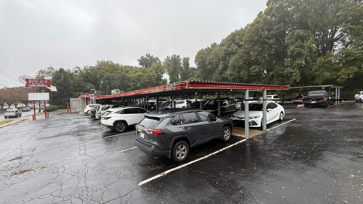A wide exterior shot of the Bar-B-Q King drive-in on a rainy day. A vintage red sign with the restaurant’s name is on the left. The parking lot is full, with cars parked under the long, covered awning that extends from the restaurant building.