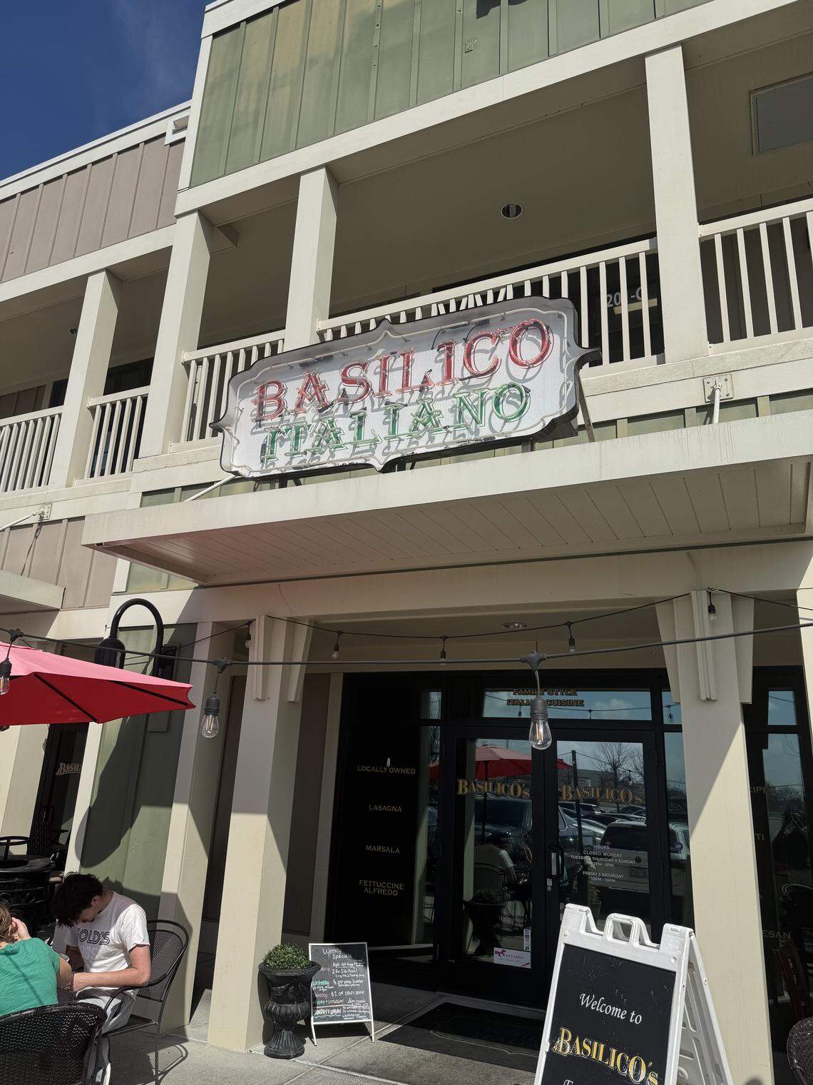 The exterior storefront of Basilico Italiano, featuring a large neon sign and an outdoor patio area with diners.