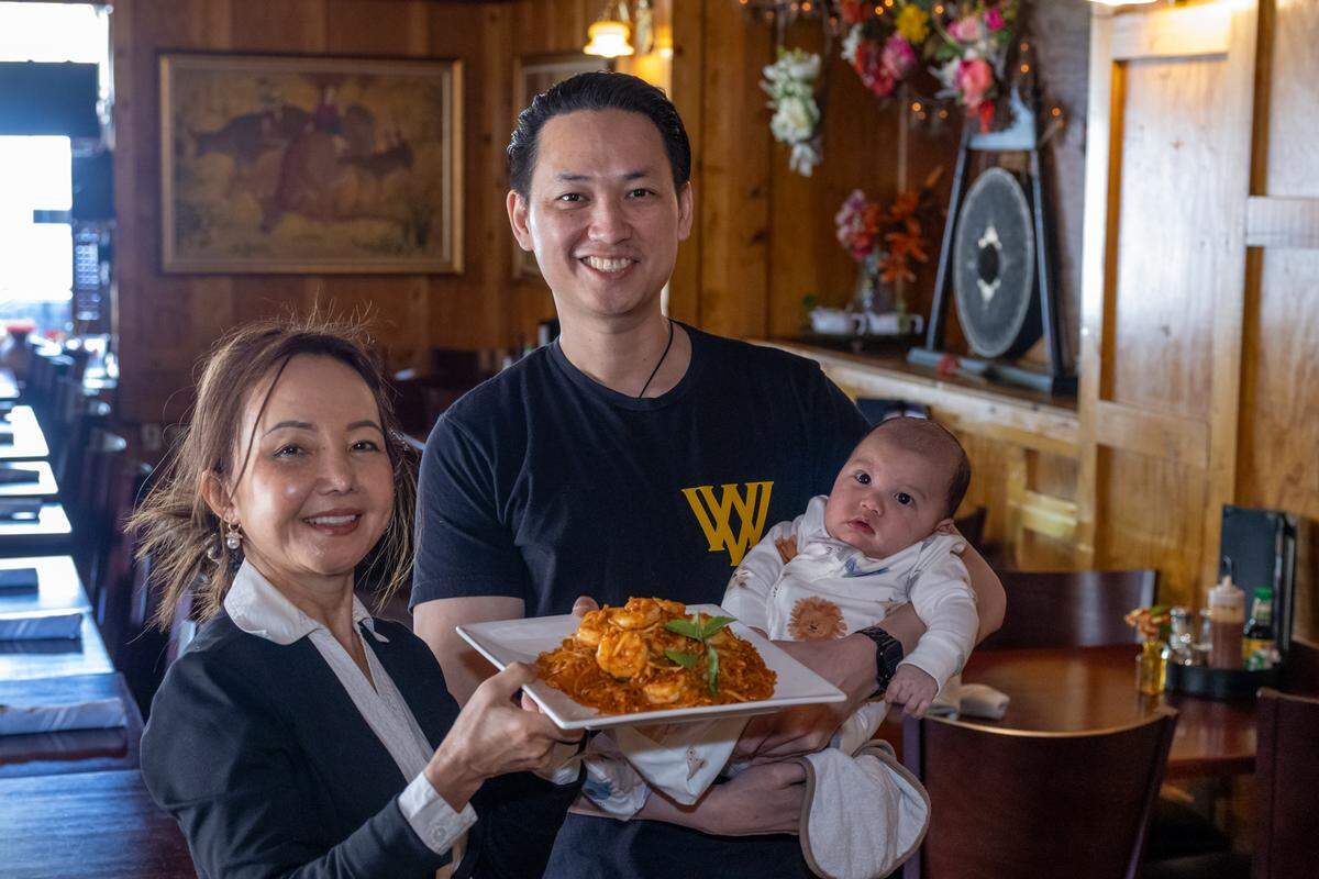 Two restaurateurs stand together smiling; one is holding a small baby and a large plate of Pad Thai. They are posing in the center of a warmly lit restaurant with traditional decor in the background.