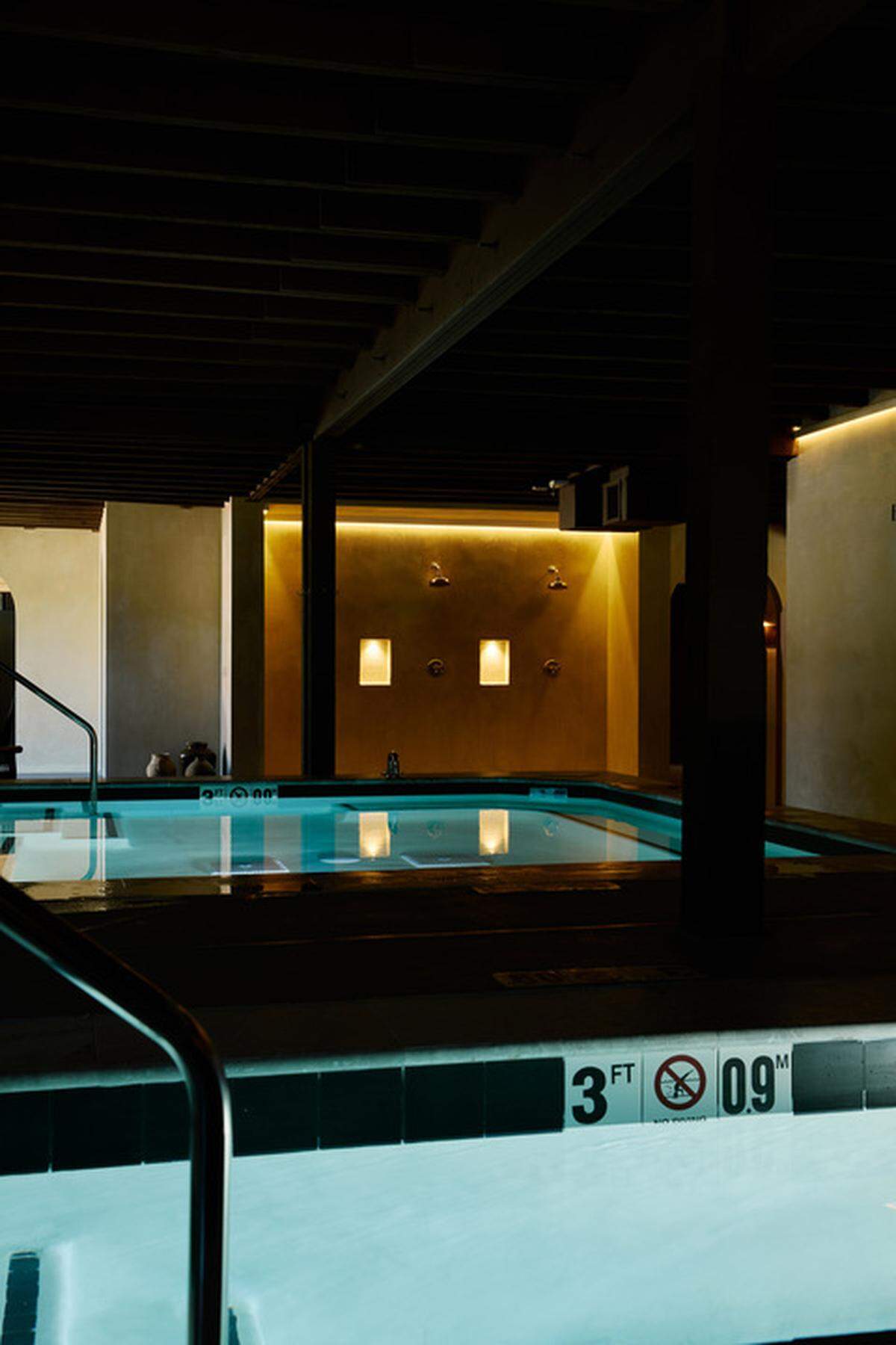 A dim, indoor swimming pool with glowing blue water. The walls are a light color and there are two shower heads with water spouts framed by warm-toned lighting. The ceiling has dark wooden beams. In the foreground, the edge of the pool is marked with a black and white tiled border showing the depth “3 FT” and “0.9 M”.