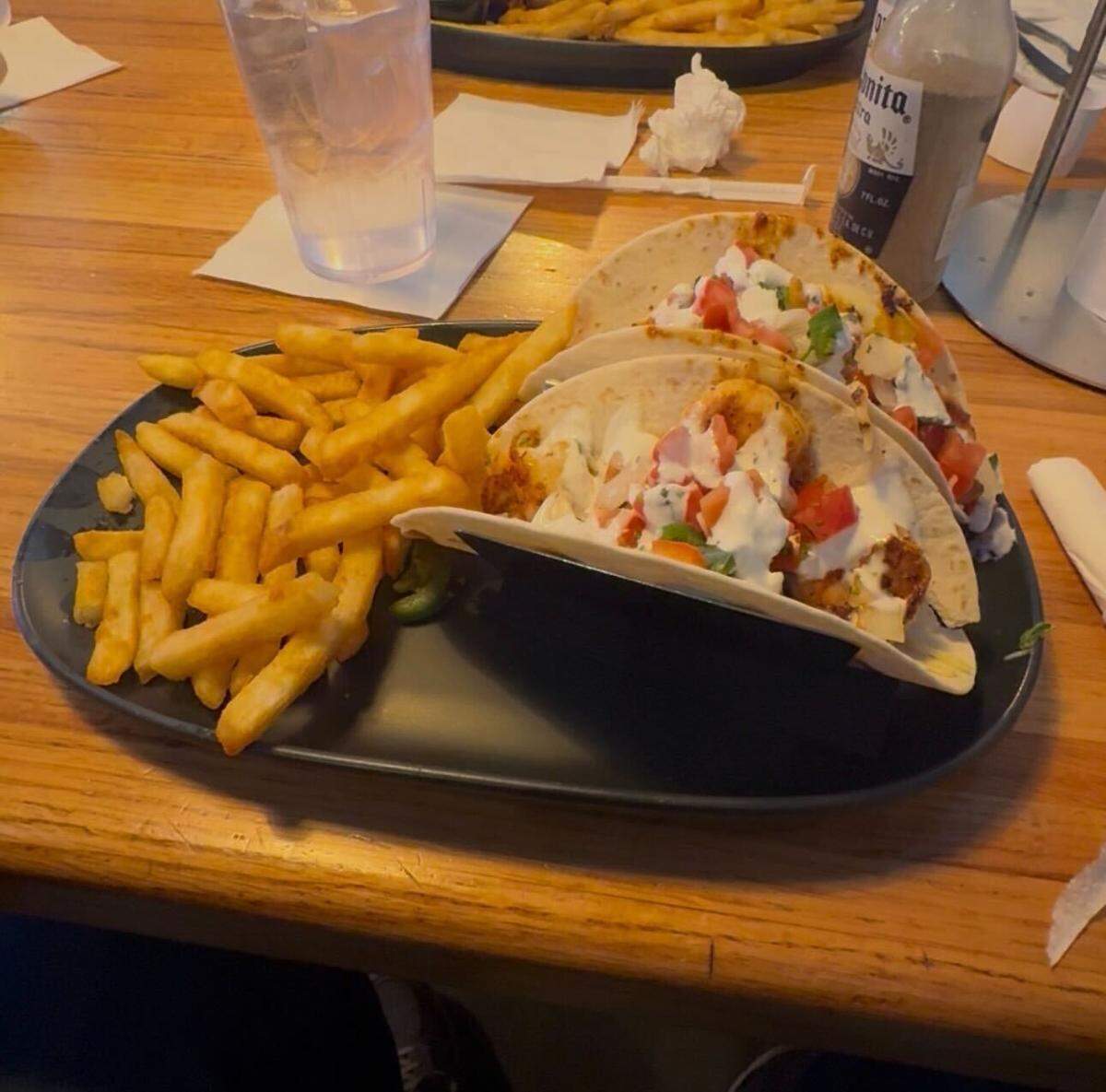 A top-down view of a meal served on a black oval plate sitting on a light-colored wooden table. The plate features two soft flour tacos filled with grilled shrimp, diced tomatoes, cilantro, and a creamy white sauce, alongside a generous side of golden-brown french fries. A glass of water and a bottle of Cholita hot sauce are visible in the background.