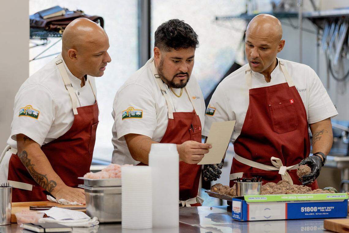 Three chefs wearing white coats and maroon aprons collaborate in a professional kitchen. The chef in the center holds and reads a slip of paper while the chef on the right, wearing black gloves, holds a large meatball over a tray. The setting includes stainless steel counters, prep containers, and kitchen equipment.