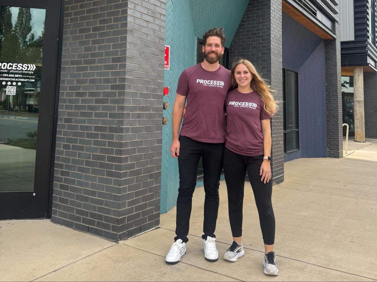 Two physical therapists stand side-by-side in matching maroon “Process Physical Therapy” t-shirts in front of their modern brick office building.
