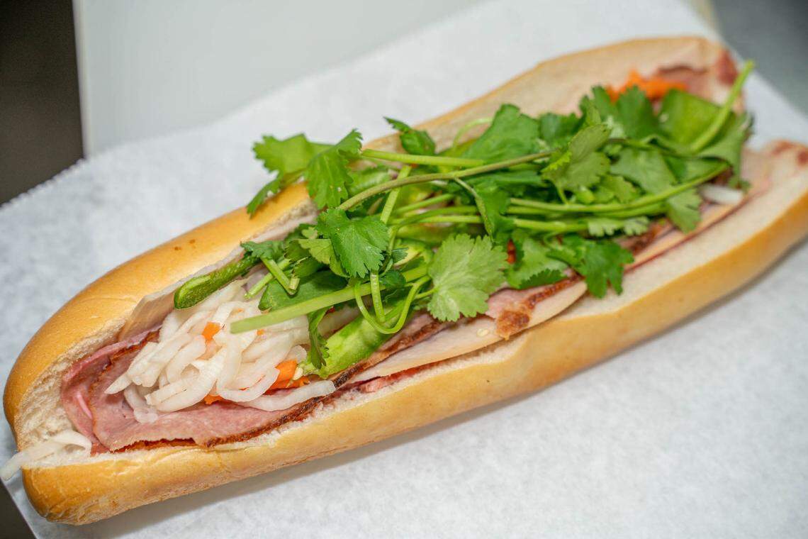 A Vietnamese bánh mì sandwich is shown on white paper. The crusty baguette is filled with layers of meat, pickled vegetables, jalapeño slices, and a large sprig of fresh cilantro.