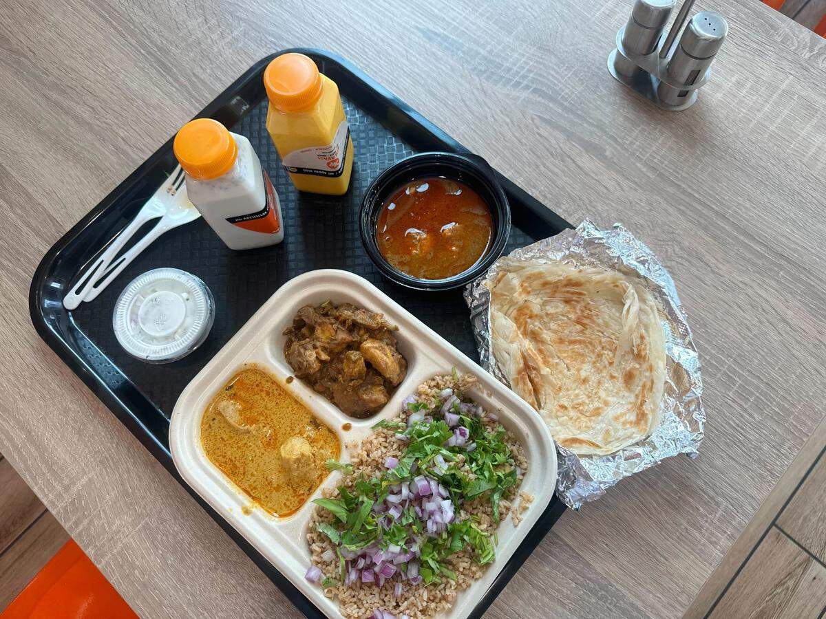 A black tray holds a variety of Indian dishes, including a three-compartment container with rice, curry, and meat, accompanied by a side of flatbread in foil. Two small bottles with orange caps and a small container of white sauce complete the meal on a light wood-textured table.