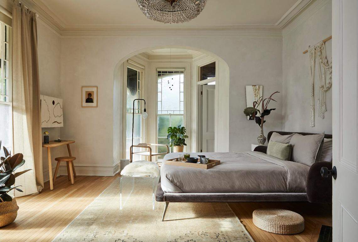A serene, light-filled bedroom suite with pale, textured walls and hardwood floors. The room features a low-profile bed covered in neutral, taupe linens, centered on a light beige area rug. The bed faces an arched alcove with large windows that overlook greenery. To the left, there is a small wooden desk, and a large, beaded chandelier hangs from the ceiling.