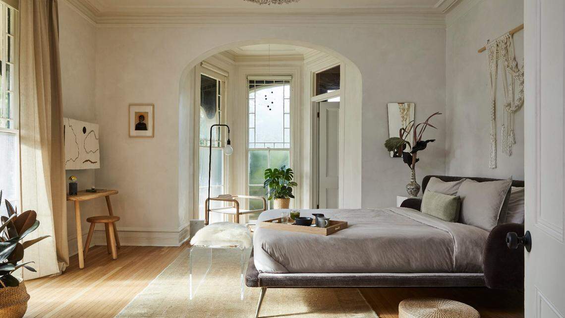 A serene, light-filled bedroom suite with pale, textured walls and hardwood floors. The room features a low-profile bed covered in neutral, taupe linens, centered on a light beige area rug. The bed faces an arched alcove with large windows that overlook greenery. To the left, there is a small wooden desk, and a large, beaded chandelier hangs from the ceiling.
