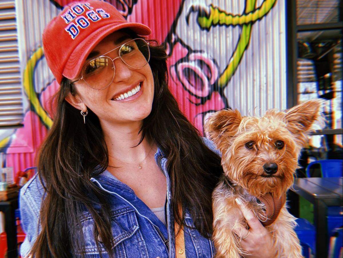 A close-up portrait of a person with long dark hair, wearing a red baseball cap that says “HOT DOGS” in white and blue lettering and wearing aviator-style glasses and a denim jacket while holding a small, brown, scruffy-haired terrier-mix dog. The background features a vibrant, colorful mural on a corrugated metal wall.