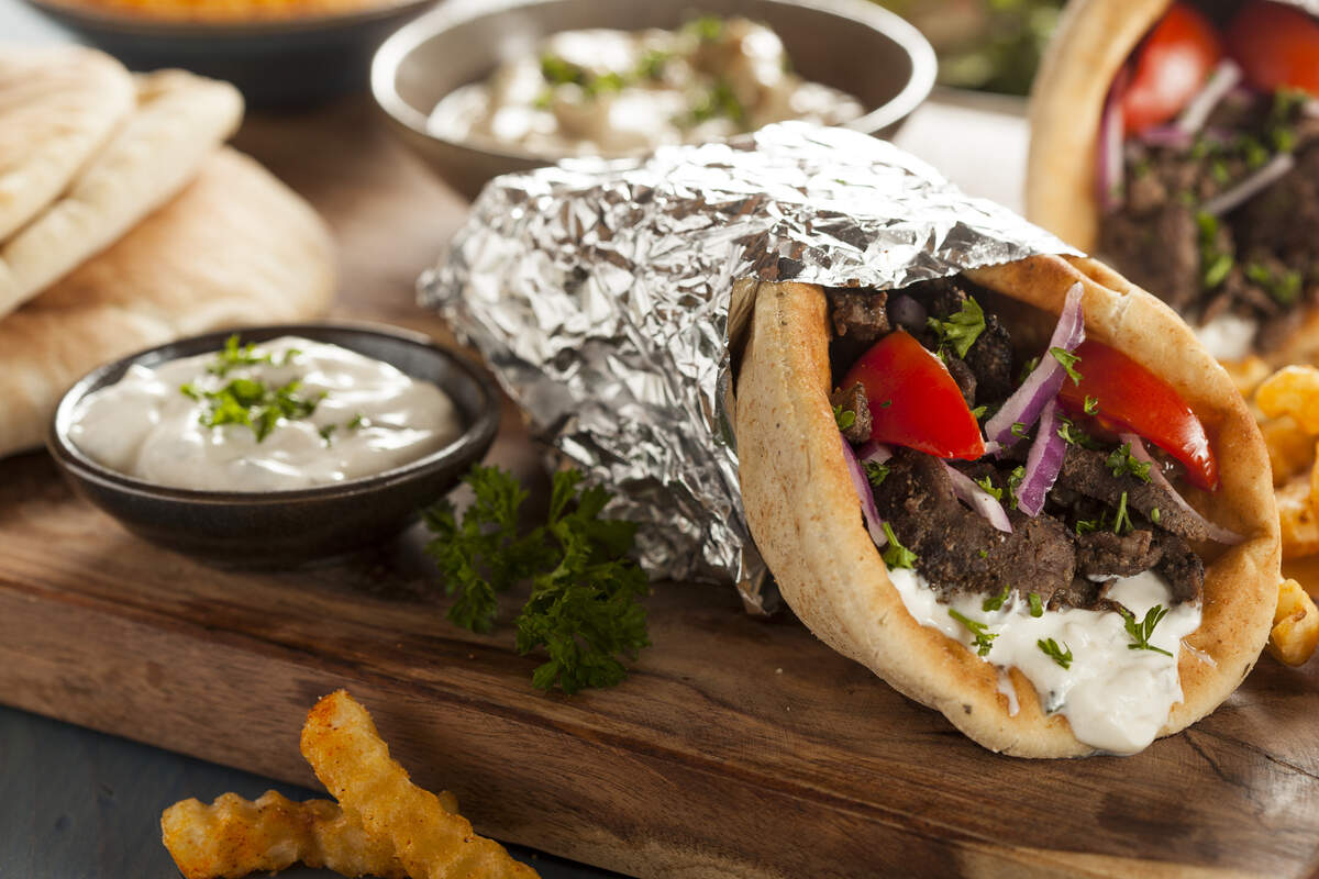 A seasoned beef gyro wrapped in warm pita bread and aluminum foil, filled with tzatziki sauce, tomatoes, onions, and parsley, served on a wooden board next to french fries and extra dipping sauce.