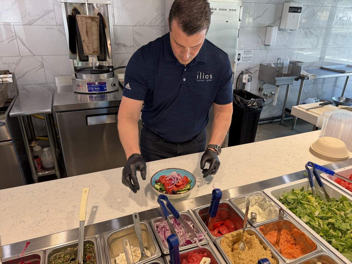 A restaurant worker wearing black gloves prepares a fresh Greek salad with tomatoes and red onions at a stainless-steel counter filled with various food service inserts.