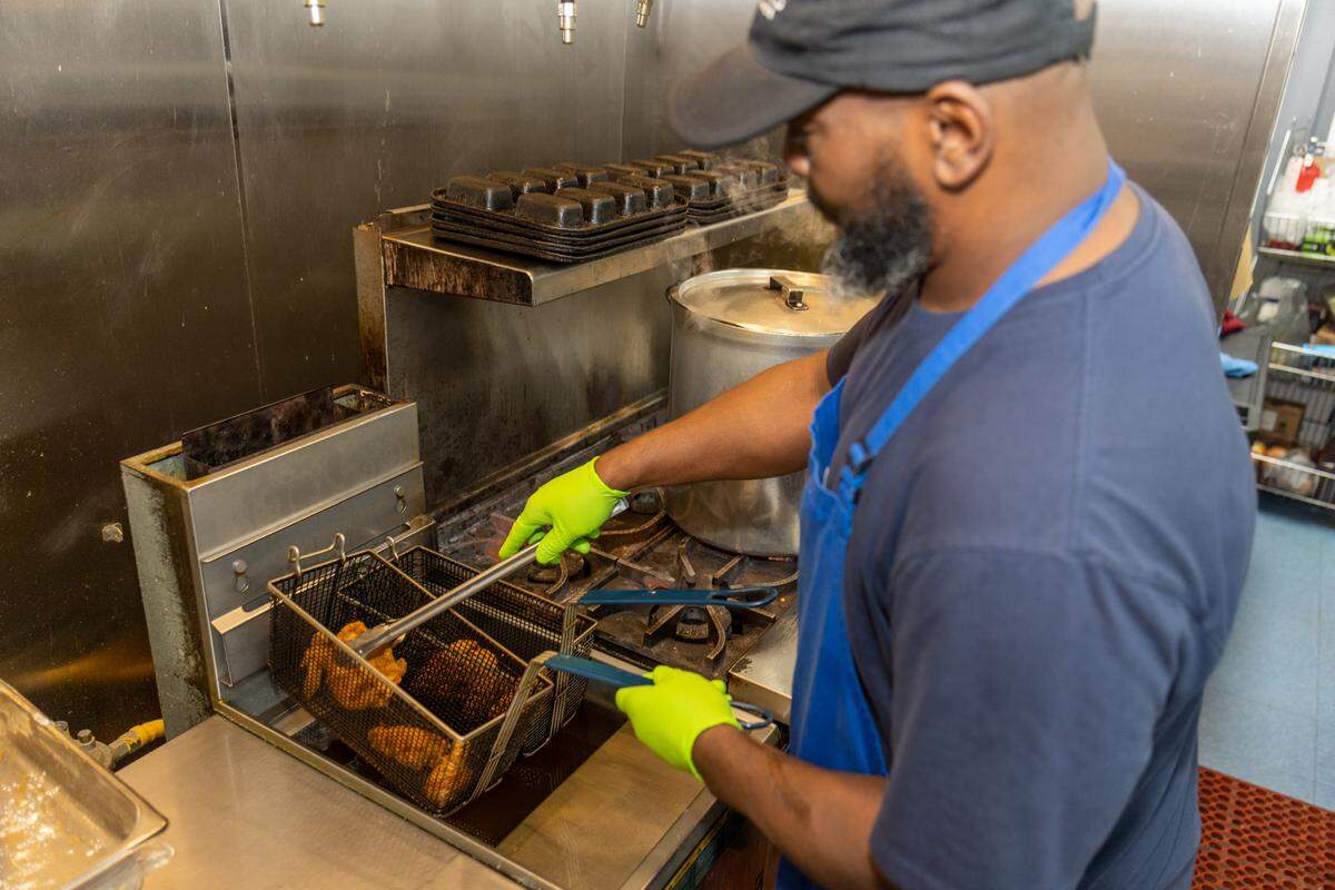 A chef wearing a navy blue shirt, a blue apron, and bright neon green gloves is using tongs to lift a frying basket filled with dark brown, fried items (likely chicken or fish) out of a deep fryer set on a stainless steel commercial stovetop. The chef has a black baseball cap and a salt-and-pepper beard. There is steam rising near a large silver pot on the stove. The kitchen area is stainless steel, and a red mat is visible on the blue floor.