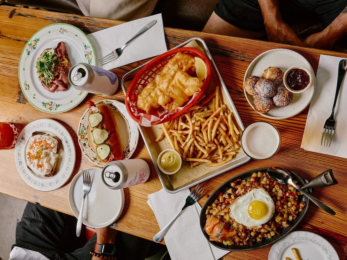 An overhead view of a wooden restaurant table filled with dishes including fish and chips, a hot dog, hash with a fried egg, and pastries.