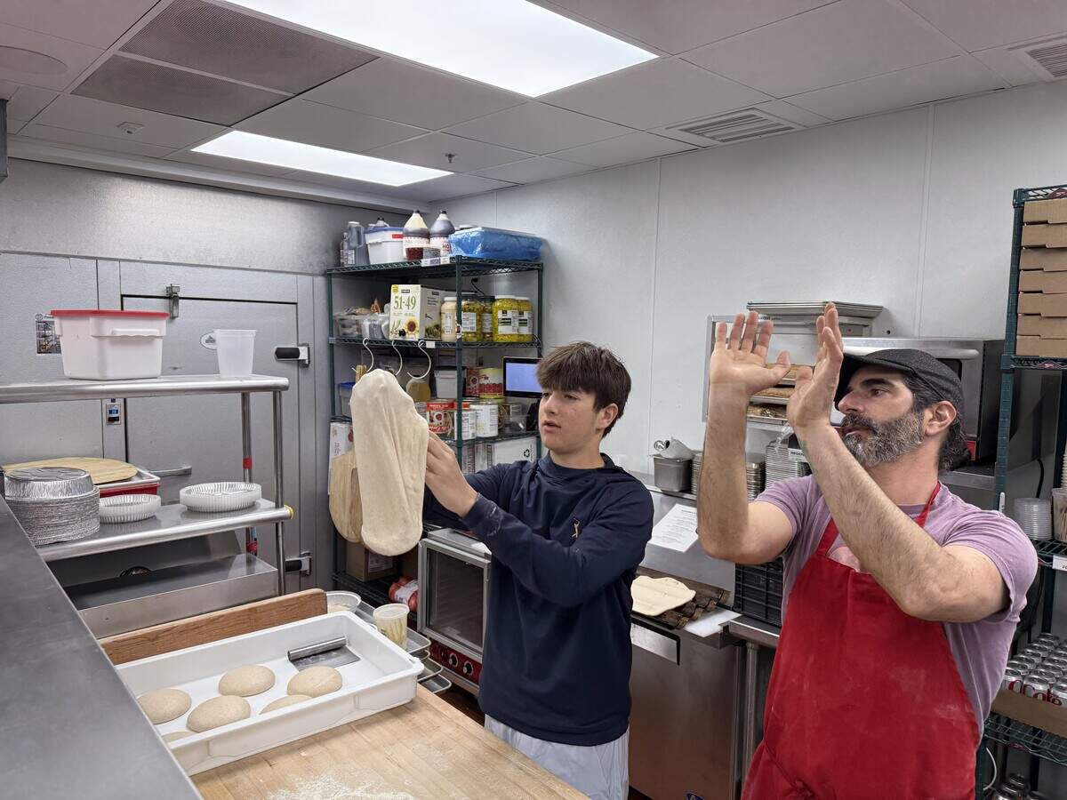 Inside a commercial kitchen, two people are making pizza. A younger person wearing a dark blue hoodie is holding up a stretched piece of pizza dough, preparing to toss it. Beside them, another person wearing a purple shirt, a red apron, and a flat cap has their hands raised in the air, appearing to guide or demonstrate the tossing technique. On the wooden counter in front of them is a white tray containing several round, smooth balls of dough.