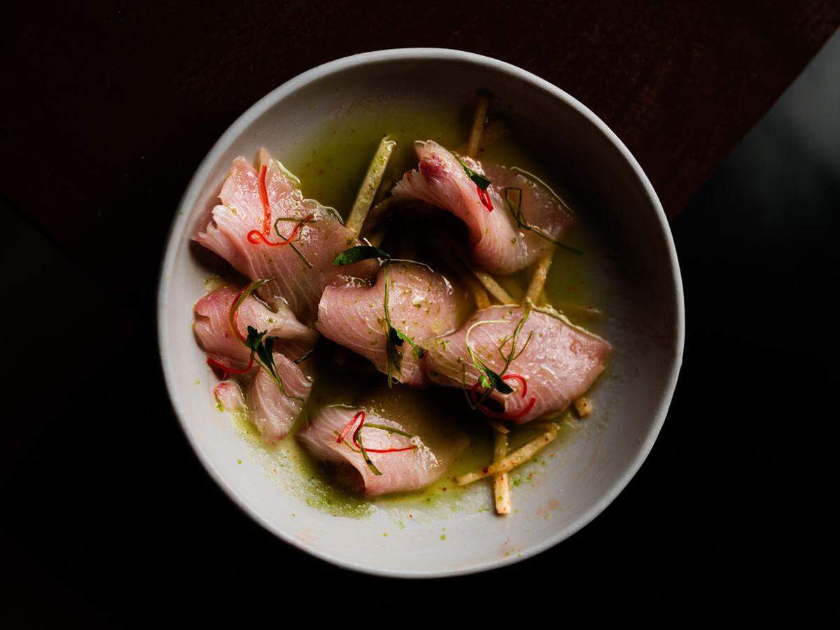 An overhead, high-contrast shot of a crudo dish served in a matte gray bowl. Thin slices of raw fish are arranged over a base of thin sticks, immersed in a light green sauce, and garnished with bright red chili strips and green microgreens.