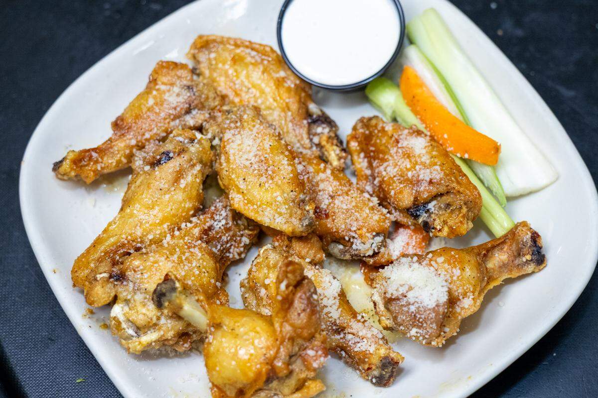 A close-up, top-down view of an appetizer plate. A generous portion of fried chicken wings is sprinkled with Parmesan cheese. On the side of the plate are several celery sticks, a slice of carrot, and a small black cup filled with a white, creamy dipping sauce.