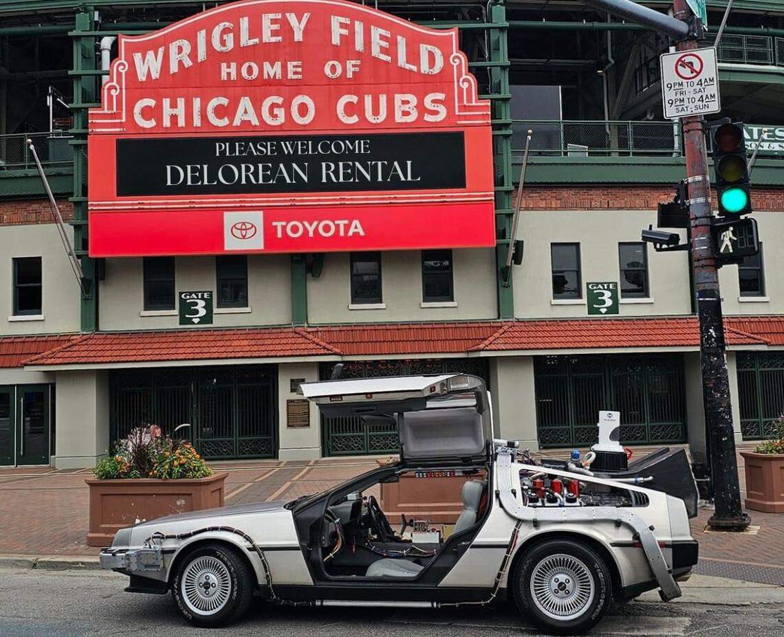 A straight-on shot of a DeLorean time machine replica parked in front of the iconic red Wrigley Field marquee. The car’s gull-wing doors are open, revealing the interior and the time machine components on its exterior. The stadium sign reads “WRIGLEY FIELD HOME OF CHICAGO CUBS,” and the digital portion below it says, “PLEASE WELCOME DELOREAN RENTAL.”