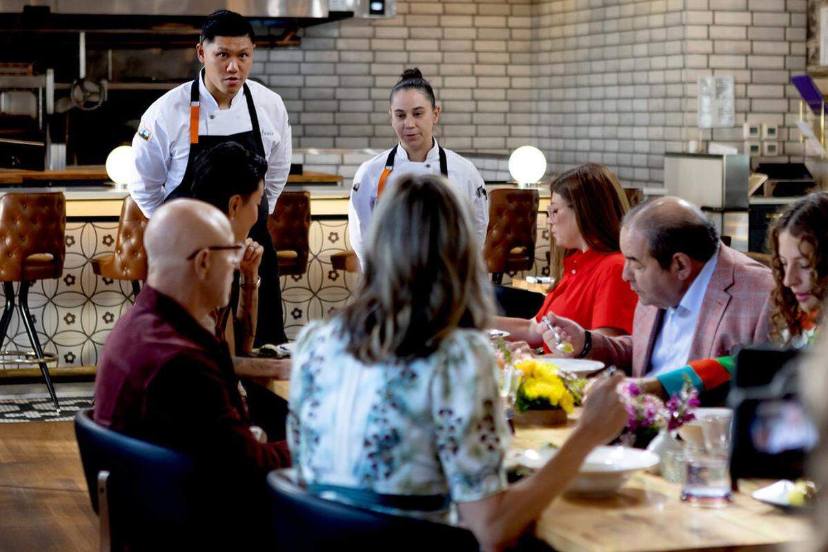 A wide shot from a high-end restaurant or culinary competition set. In the foreground, several guests sit at a long wooden table decorated with bright yellow and purple flowers, partially obscured from the back. In the background, two chefs in white coats and black aprons stand attentively near a tiled bar with tufted leather stools, appearing to present a dish or listen to feedback. The modern space features white brick walls and warm, ambient lighting.